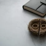Two gold wedding bands resting on a split nest of straw on a marble desk, beside a leather notebook and pen, symbolising superannuation division during divorce
